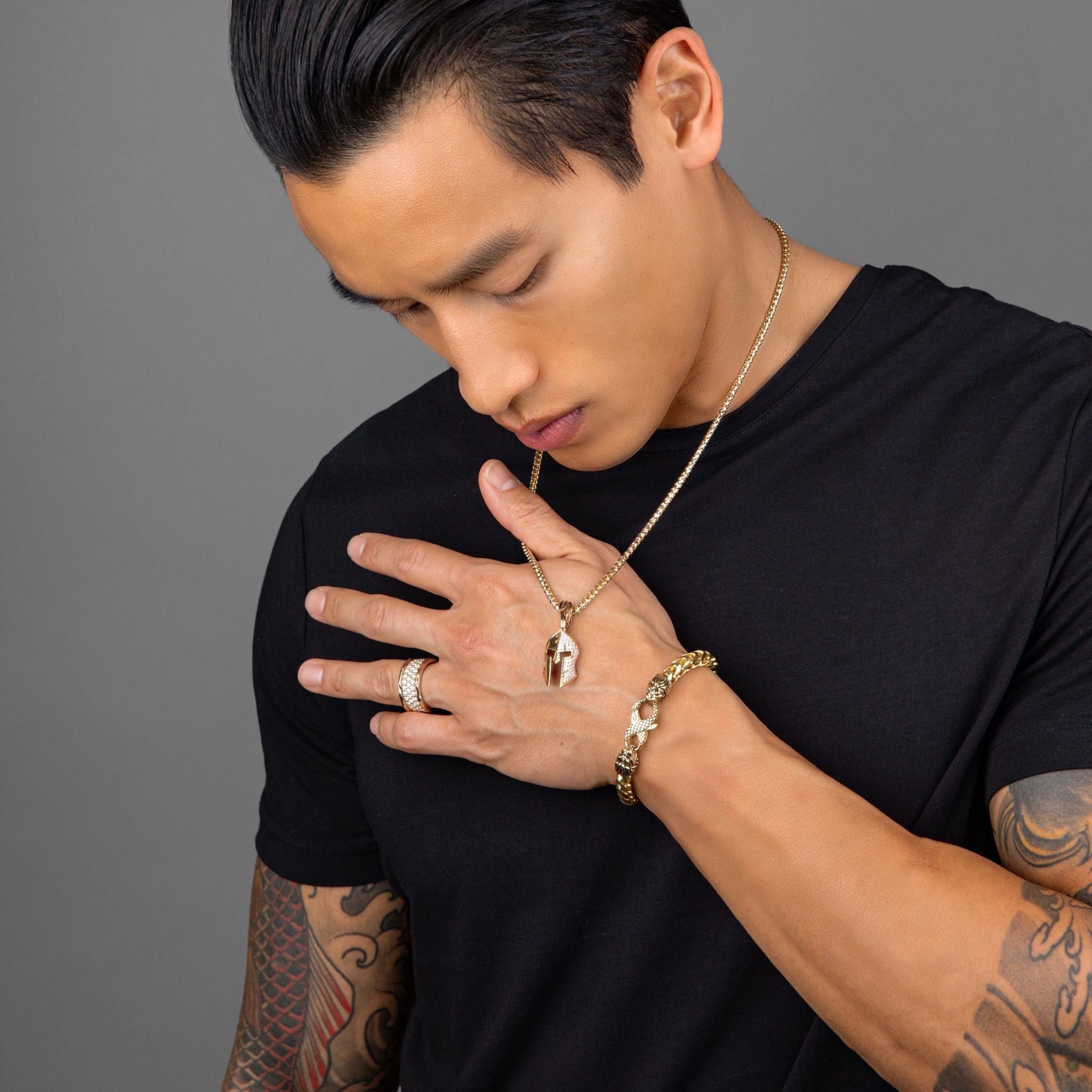 a tattooed man holding up a gold Spartan necklace that is covered with diamonds on one half of the helmet