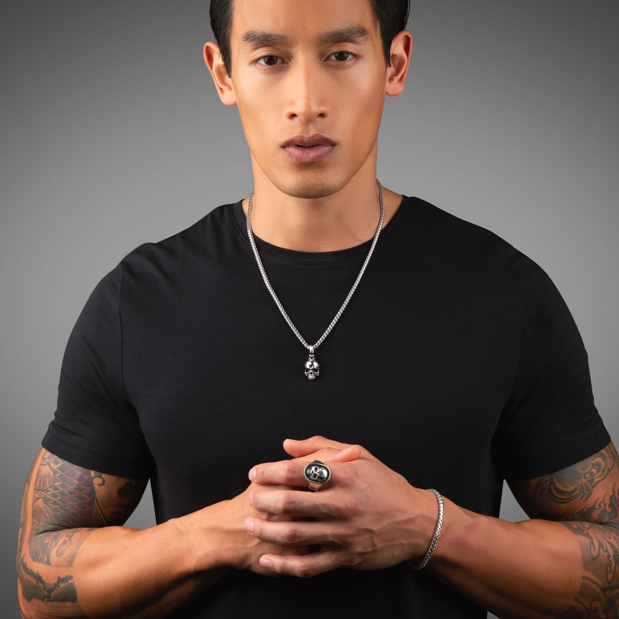 a man in a black shirt stands with his hands hands folded, showing a silver skull ring and a skull pendant